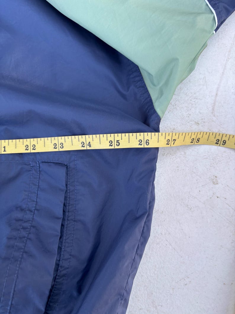 May include: A navy blue and light green jacket with a white trim. The jacket is being measured with a yellow measuring tape. The jacket has a pocket on the front. The jacket is made of a lightweight material.
