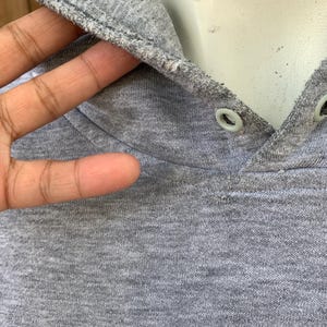 May include: Close-up of a gray hoodie with a drawstring hood. The hood is lined with a lighter gray fabric and has two eyelets. The fabric appears to be a soft, knit material.