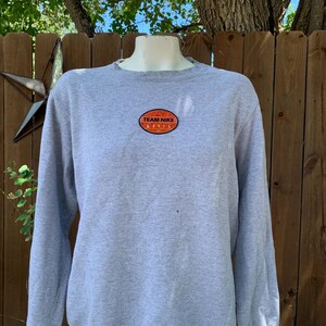 May include: A light gray sweatshirt with long sleeves. The sweatshirt features a small, oval patch on the chest with the words "TEAM NIKE" in orange and white. The background includes a wooden fence and foliage.