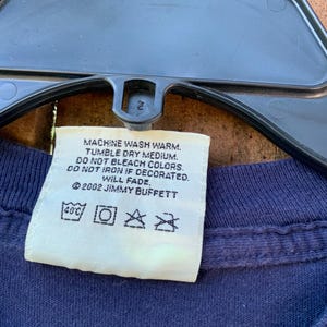 May include: White care label with instructions for washing a garment. The label says "Machine wash warm. Tumble dry medium. Do not bleach colors. Do not iron if decorated. Will fade. &copy;2002 Jimmy Buffett". The label also has symbols for washing temperature, machine wash, tumble dry, and do not iron.