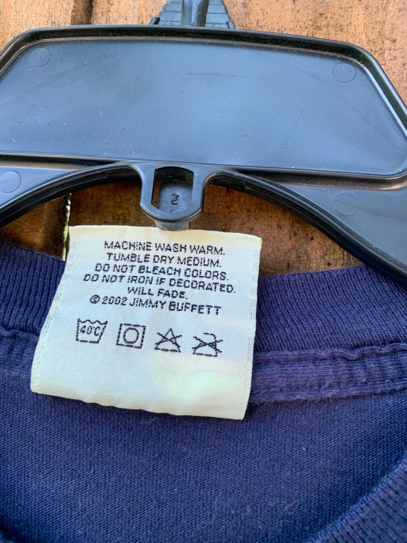 May include: White care label with instructions for washing a garment. The label says "Machine wash warm. Tumble dry medium. Do not bleach colors. Do not iron if decorated. Will fade. &copy;2002 Jimmy Buffett". The label also has symbols for washing temperature, machine wash, tumble dry, and do not iron.