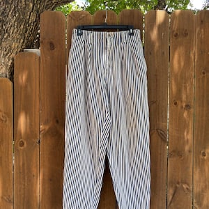 Puede incluir: Pantalones de cintura alta a rayas blancas y azul marino colgados de una percha negra contra una valla de madera. Los pantalones tienen una parte delantera plisada y cierre de bot&oacute;n. Las rayas verticales crean un look cl&aacute;sico y n&aacute;utico.