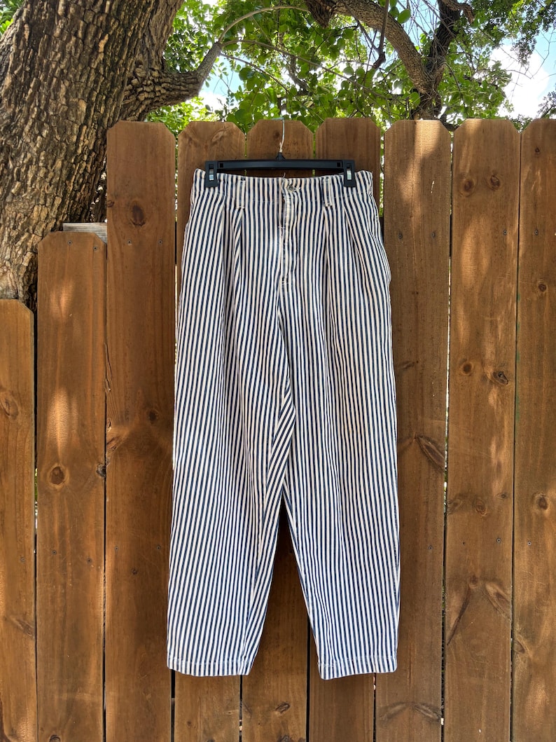 Puede incluir: Pantalones de cintura alta a rayas blancas y azul marino colgados de una percha negra contra una valla de madera. Los pantalones tienen una parte delantera plisada y cierre de bot&oacute;n. Las rayas verticales crean un look cl&aacute;sico y n&aacute;utico.