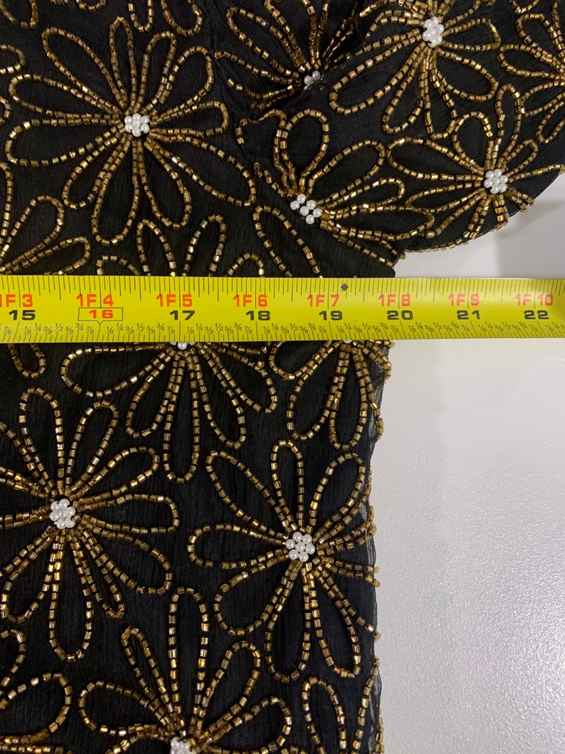 May include: Black sheer fabric with gold and white beading in a floral pattern. The fabric is measured with a tape measure.