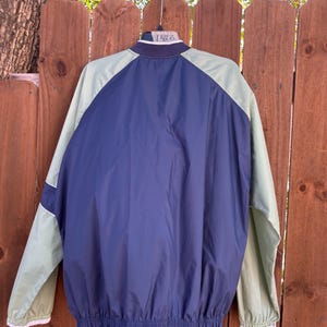 May include: A navy blue and light green jacket with white trim. The jacket has a navy blue body with light green sleeves and shoulders. The jacket is hanging on a hanger against a wooden fence.