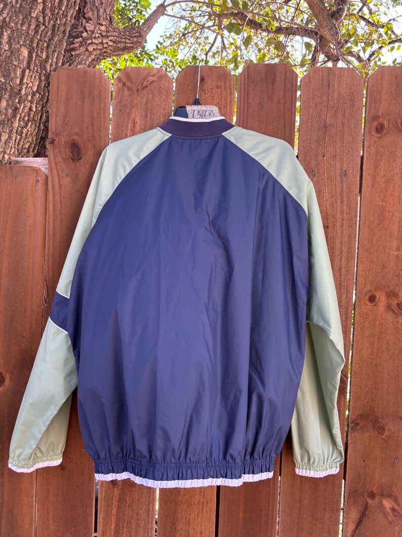 May include: A navy blue and light green jacket with white trim. The jacket has a navy blue body with light green sleeves and shoulders. The jacket is hanging on a hanger against a wooden fence.