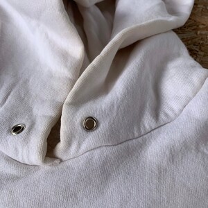May include: Close-up of a white hooded sweatshirt with two metal eyelets on the hood.