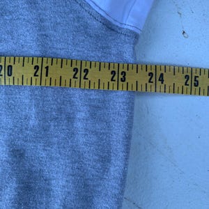 Puede incluir: Una prenda gris y blanca se mide con una cinta m&eacute;trica amarilla. La cinta m&eacute;trica muestra una medida entre 50,8 y 63,5 cm. La prenda parece estar hecha de una tela de punto gris con tela blanca en el hombro.
