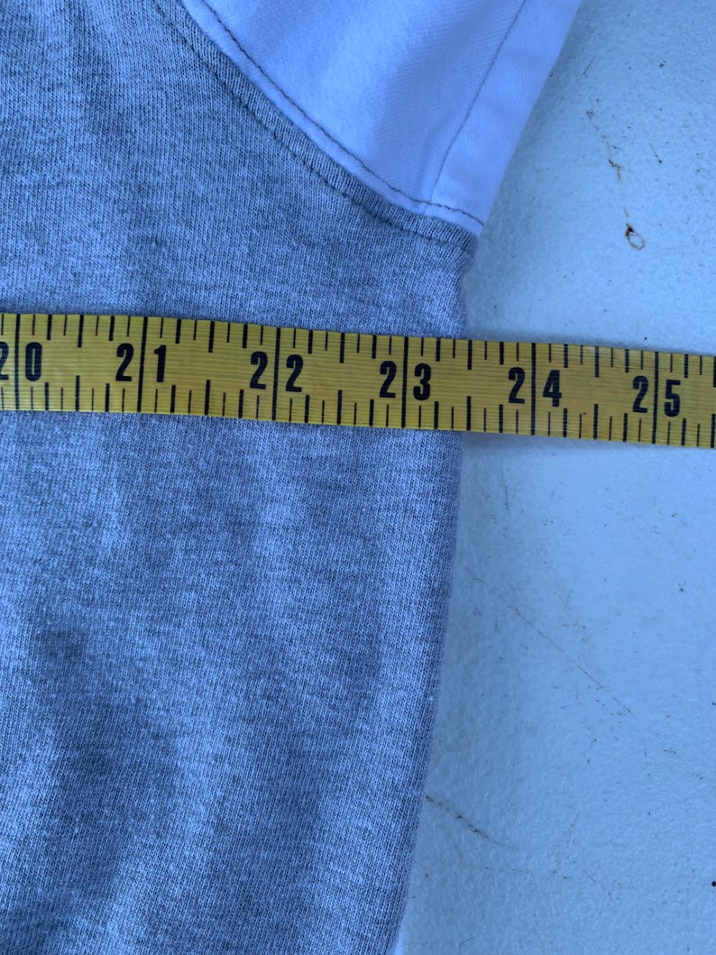 Puede incluir: Una prenda gris y blanca se mide con una cinta m&eacute;trica amarilla. La cinta m&eacute;trica muestra una medida entre 50,8 y 63,5 cm. La prenda parece estar hecha de una tela de punto gris con tela blanca en el hombro.