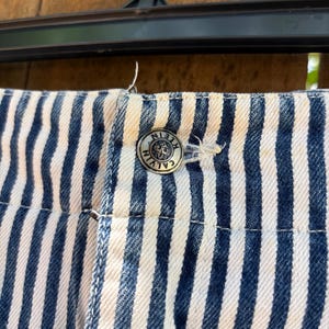 Puede incluir: Primer plano de tela vaquera a rayas azules y blancas. Se ve un bot&oacute;n plateado con el logotipo de Calvin Klein. La tela forma parte de un pantal&oacute;n corto o pantal&oacute;n.