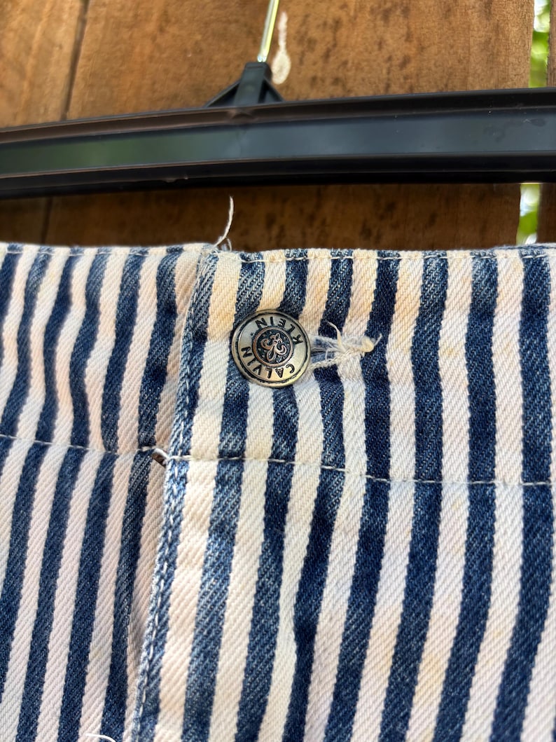 Puede incluir: Primer plano de tela vaquera a rayas azules y blancas. Se ve un bot&oacute;n plateado con el logotipo de Calvin Klein. La tela forma parte de un pantal&oacute;n corto o pantal&oacute;n.