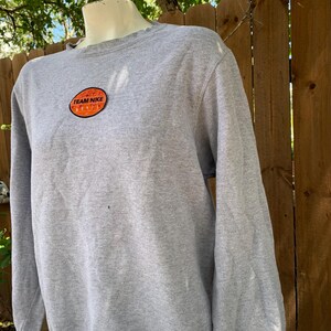 May include: A heather gray sweatshirt with long sleeves. The sweatshirt features a circular patch with the words "TEAM NIKE" in white lettering on an orange background. The sweatshirt is displayed on a mannequin.