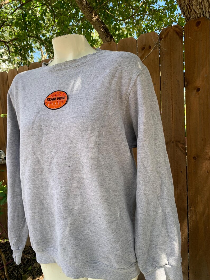 May include: A heather gray sweatshirt with long sleeves. The sweatshirt features a circular patch with the words "TEAM NIKE" in white lettering on an orange background. The sweatshirt is displayed on a mannequin.