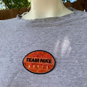 May include: A gray crew neck t-shirt with a Team Nike patch. The patch is oval-shaped with an orange background, black border, and the words "TEAM NIKE" in white. The patch also features running figures.