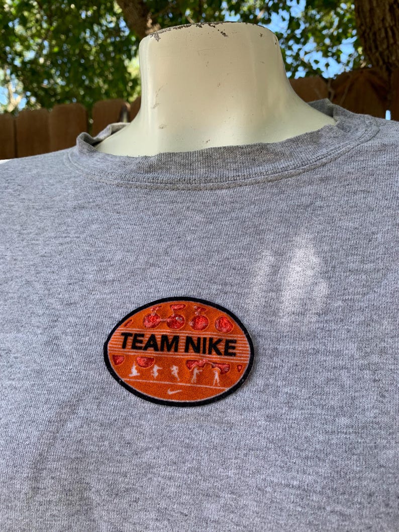May include: A gray crew neck t-shirt with a Team Nike patch. The patch is oval-shaped with an orange background, black border, and the words "TEAM NIKE" in white. The patch also features running figures.