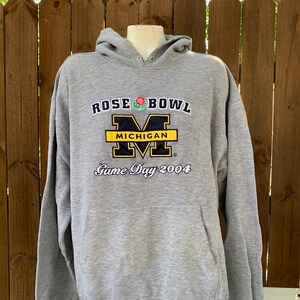 May include: A gray hooded sweatshirt with the text "ROSE BOWL MICHIGAN Game Day 2004" in navy blue, yellow, and white. The sweatshirt has a front pocket and a drawstring hood. The logo features a rose and the letter "M".