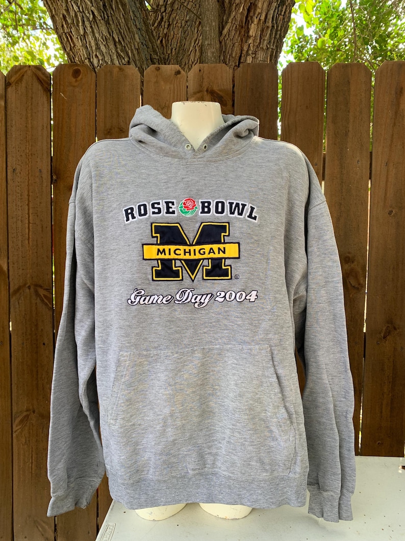 May include: A gray hooded sweatshirt with the text "ROSE BOWL MICHIGAN Game Day 2004" in navy blue, yellow, and white. The sweatshirt has a front pocket and a drawstring hood. The logo features a rose and the letter "M".