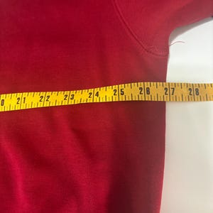 May include: A close-up of a red garment with a yellow measuring tape laid across it. The tape shows measurements from 20 to 28 inches. The garment appears to be made of a soft, woven material.