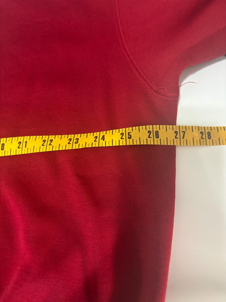 May include: A close-up of a red garment with a yellow measuring tape laid across it. The tape shows measurements from 20 to 28 inches. The garment appears to be made of a soft, woven material.