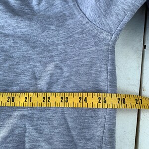 May include: A light gray t-shirt with a crew neck. A yellow measuring tape is laid across the fabric, showing measurements from 20 to 27 inches.