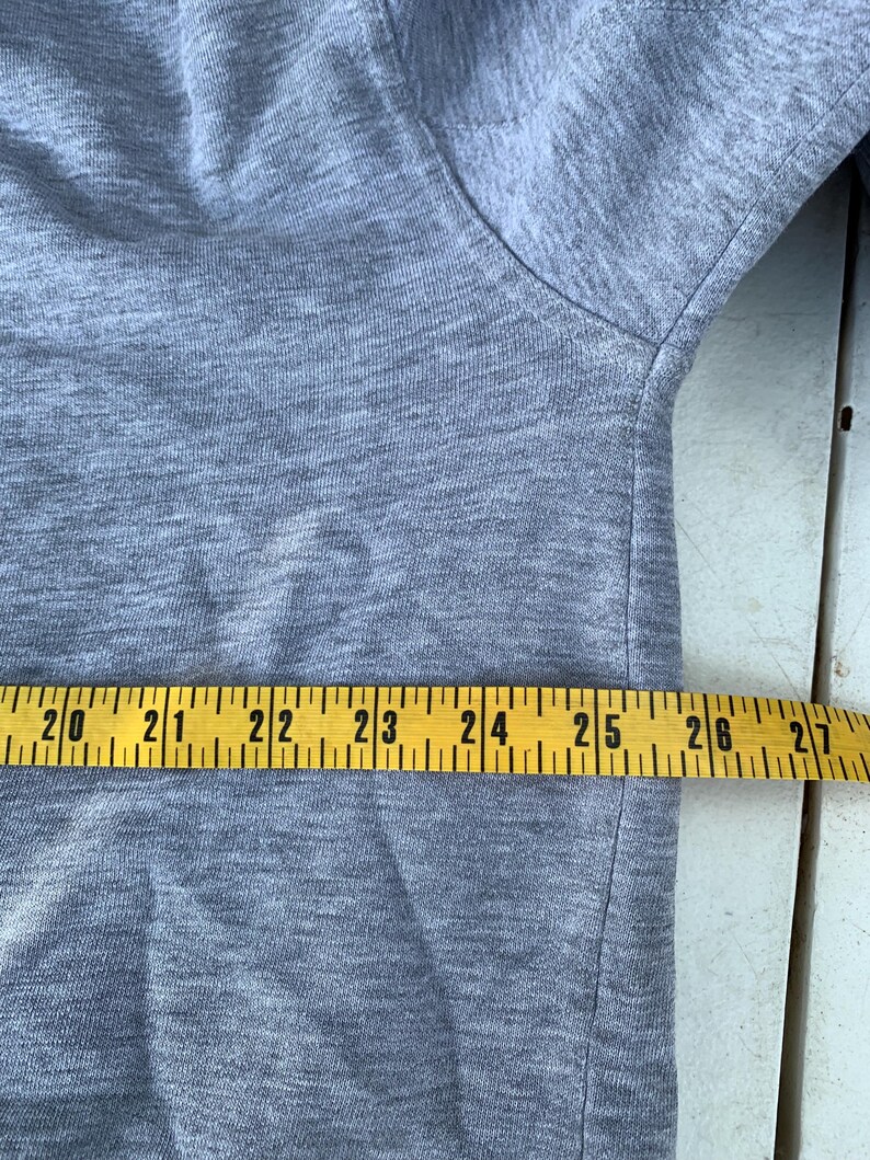 May include: A light gray t-shirt with a crew neck. A yellow measuring tape is laid across the fabric, showing measurements from 20 to 27 inches.