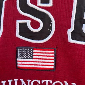 May include: Close-up of a maroon sweatshirt with the word "USA" in large, black, white-outlined letters. A small embroidered American flag patch is centered below the letters. The word "HINGTON" is embroidered in white below the flag.