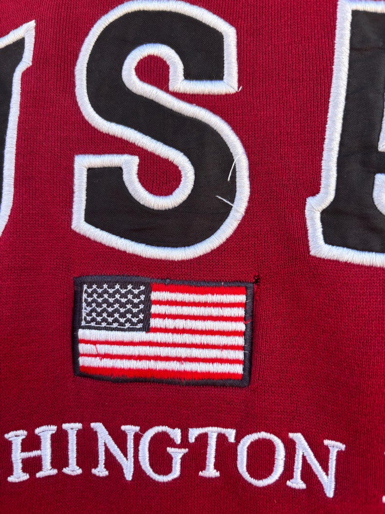 May include: Close-up of a maroon sweatshirt with the word "USA" in large, black, white-outlined letters. A small embroidered American flag patch is centered below the letters. The word "HINGTON" is embroidered in white below the flag.