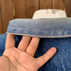 May include: Close-up of a blue denim jacket collar with a white collar support. The denim has a classic wash and visible stitching. The jacket is on a white form, with a wooden fence in the background.