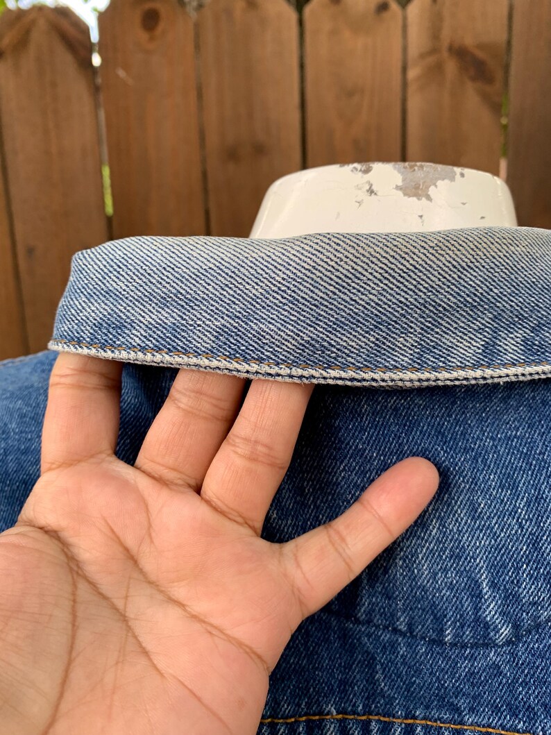 May include: Close-up of a blue denim jacket collar with a white collar support. The denim has a classic wash and visible stitching. The jacket is on a white form, with a wooden fence in the background.