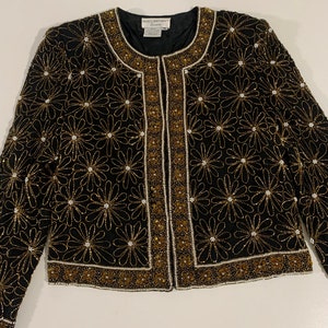 May include: A black beaded jacket with gold and white accents. The jacket features a floral pattern and a front zipper closure.