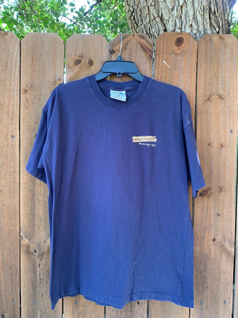 May include: A navy blue t-shirt with a graphic of a wooden sign that reads "Margaritaville Montego Bay".