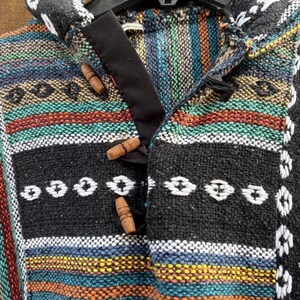 Pode incluir: Um poncho tecido colorido com capuz, com um padr&atilde;o listrado em tons de preto, azul, laranja e verde. O poncho tem fechos de altern&acirc;ncia de madeira e &eacute; exibido em um cabide preto. O design inclui detalhes bordados em branco.