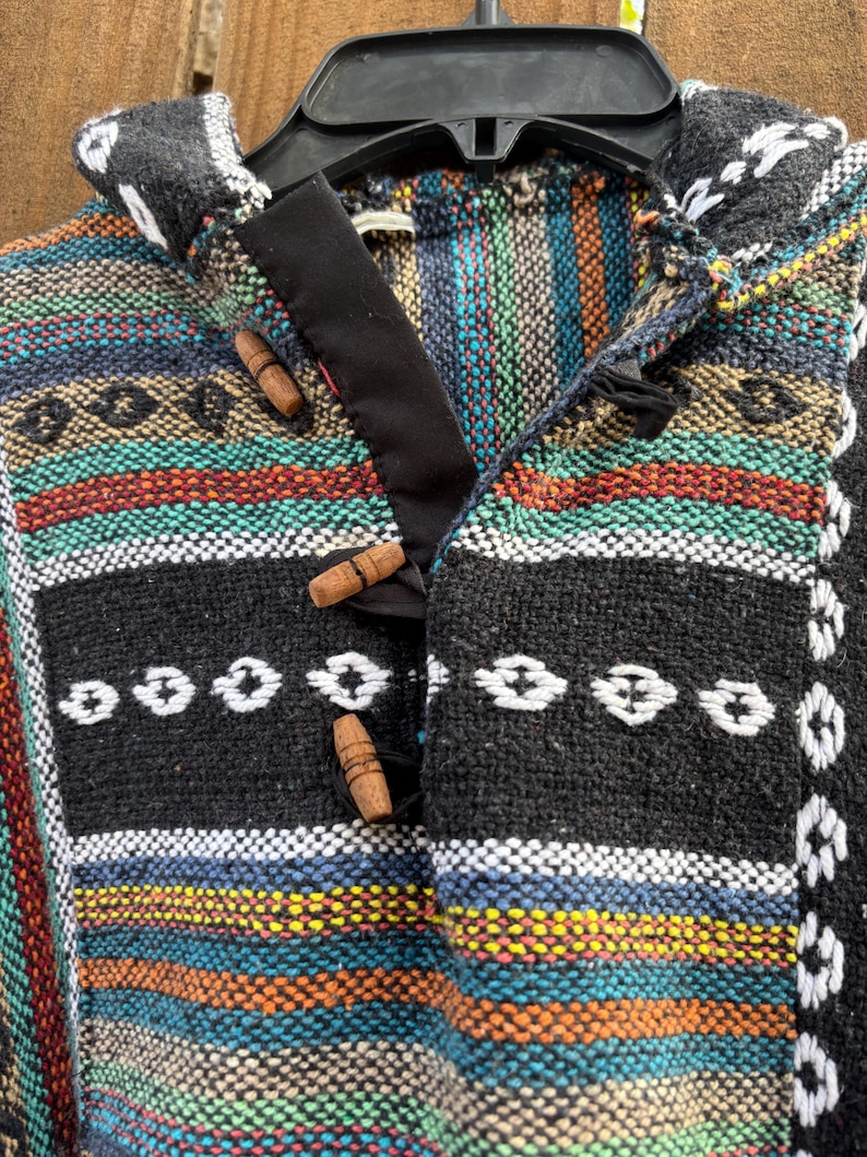 Pode incluir: Um poncho tecido colorido com capuz, com um padr&atilde;o listrado em tons de preto, azul, laranja e verde. O poncho tem fechos de altern&acirc;ncia de madeira e &eacute; exibido em um cabide preto. O design inclui detalhes bordados em branco.
