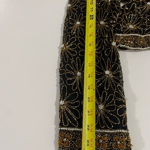 May include: A black sheer top with a floral pattern made of gold and white beads. The top has a long sleeve and a beaded trim around the cuffs.