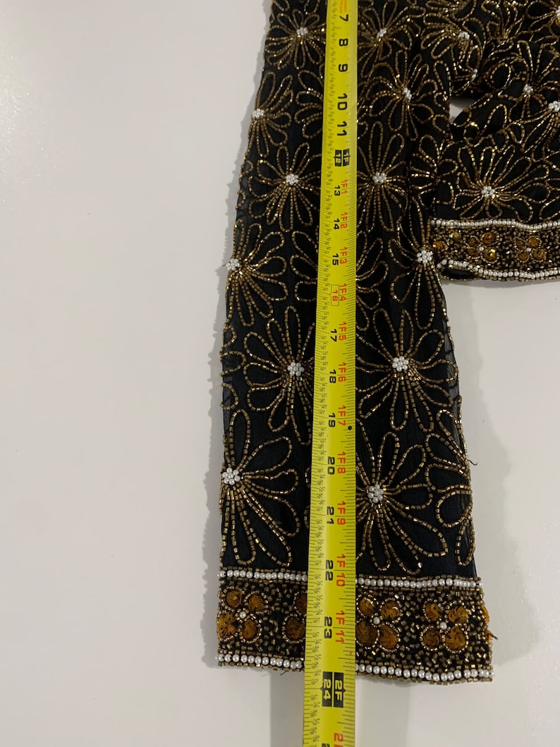 May include: A black sheer top with a floral pattern made of gold and white beads. The top has a long sleeve and a beaded trim around the cuffs.