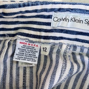 Puede incluir: Primer plano de unos pantalones cortos vaqueros a rayas azules y blancas. Los pantalones cortos tienen una etiqueta que dice "MADE IN U.S.A." y "100% COTTON". La etiqueta de talla dice "12". La etiqueta de Calvin Klein Sportswear es visible.
