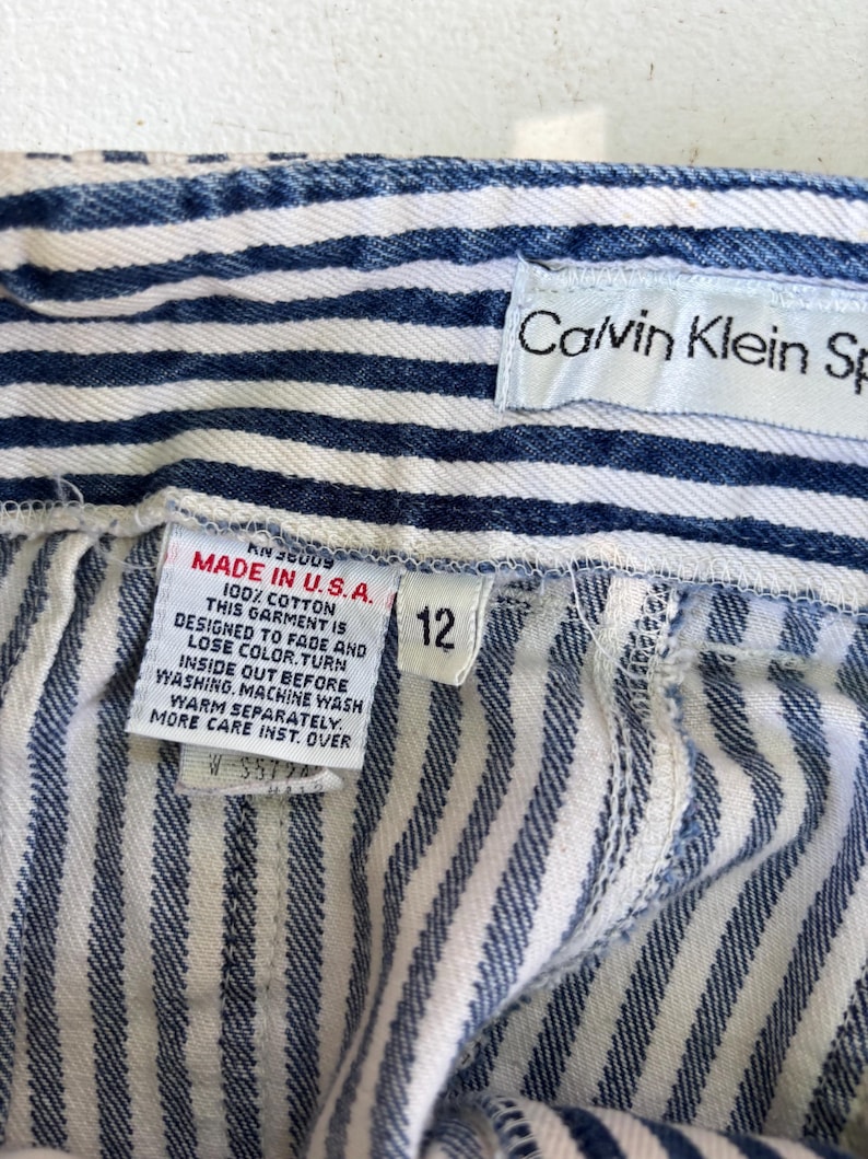 Puede incluir: Primer plano de unos pantalones cortos vaqueros a rayas azules y blancas. Los pantalones cortos tienen una etiqueta que dice "MADE IN U.S.A." y "100% COTTON". La etiqueta de talla dice "12". La etiqueta de Calvin Klein Sportswear es visible.