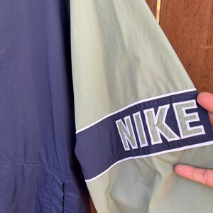 May include: A close-up of a navy blue and light green Nike jacket. The jacket features a navy blue stripe with the word "NIKE" in white block letters on the sleeve. The jacket is made of a lightweight material.