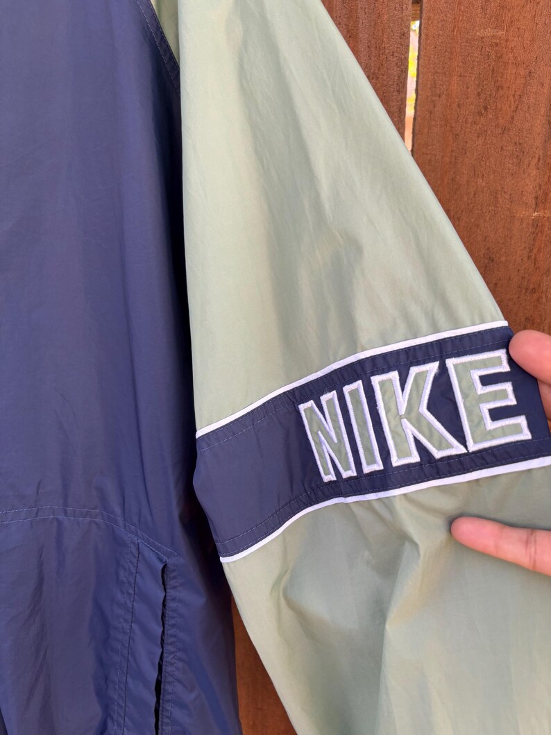 May include: A close-up of a navy blue and light green Nike jacket. The jacket features a navy blue stripe with the word "NIKE" in white block letters on the sleeve. The jacket is made of a lightweight material.