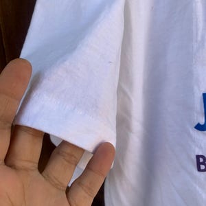 Pu&ograve; includere: Primo piano di una manica di una maglietta bianca. La manica &egrave; realizzata in un tessuto morbido e leggermente stropicciato. Le lettere "J" e "B" sono visibili in blu e viola, rispettivamente, sulla maglietta.