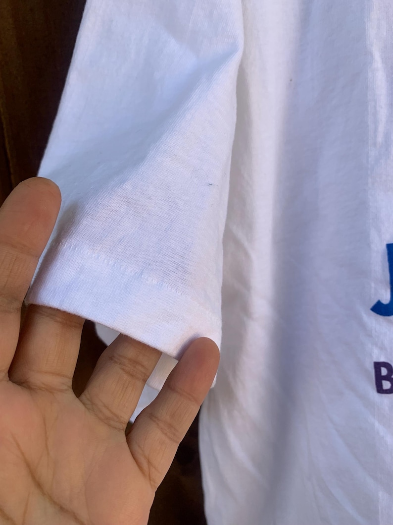 Pu&ograve; includere: Primo piano di una manica di una maglietta bianca. La manica &egrave; realizzata in un tessuto morbido e leggermente stropicciato. Le lettere "J" e "B" sono visibili in blu e viola, rispettivamente, sulla maglietta.