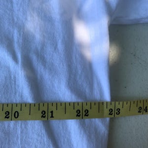 Pu&ograve; includere: Una maglietta bianca con un metro a nastro giallo appoggiato sopra. Il nastro misura da 50,8 cm a 61 cm. Il tessuto ha una trama a coste.
