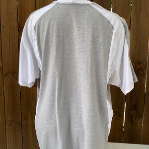 Puede incluir: Una camiseta gris con detalles blancos en los hombros y las mangas. Se ve la parte trasera de la camiseta, mostrando los colores contrastantes y el dise&ntilde;o. La camiseta se exhibe en un maniqu&iacute;.