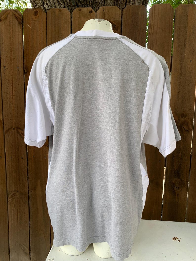 Puede incluir: Una camiseta gris con detalles blancos en los hombros y las mangas. Se ve la parte trasera de la camiseta, mostrando los colores contrastantes y el dise&ntilde;o. La camiseta se exhibe en un maniqu&iacute;.