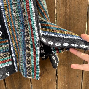 Pode incluir: Um colorido moletom Baja tecido com listras verticais em verde-azulado, vermelho, amarelo e azul. O moletom tem uma borda preta com costuras brancas e um bolso. O tecido parece grosso e texturizado.
