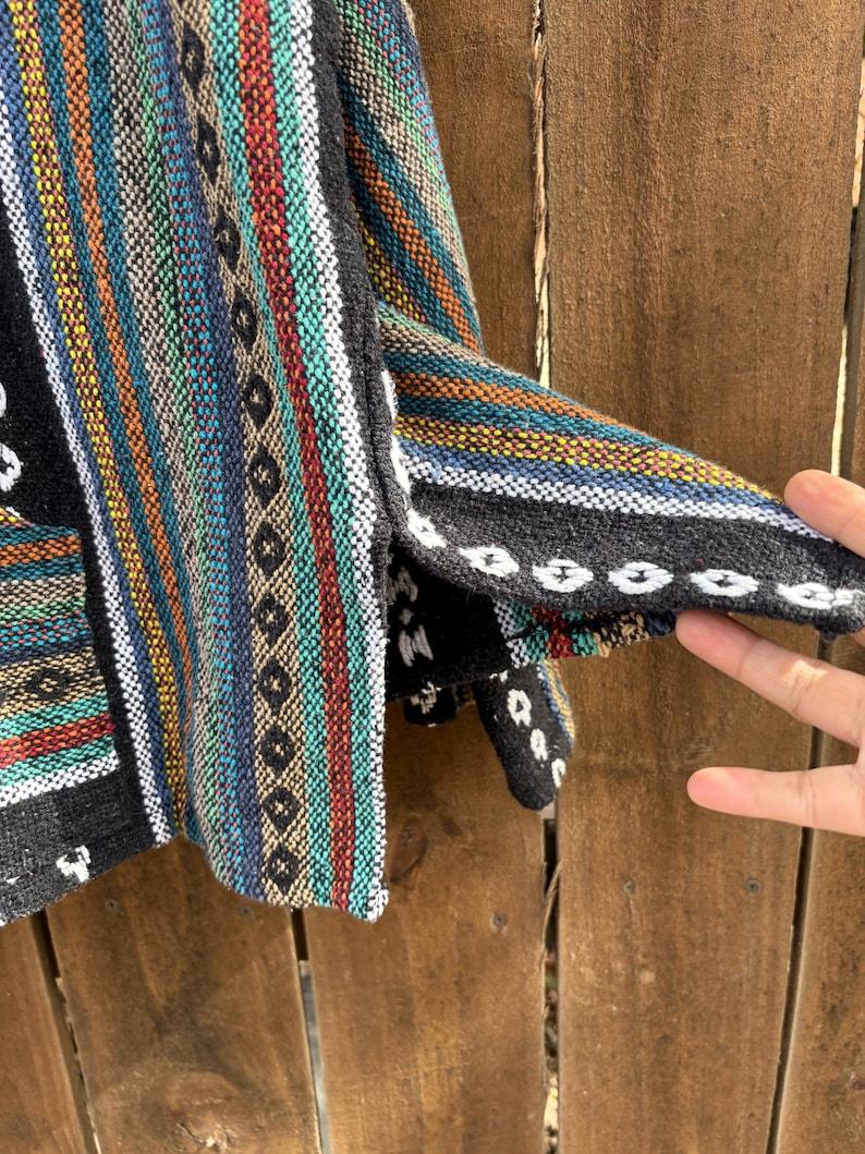 Pode incluir: Um colorido moletom Baja tecido com listras verticais em verde-azulado, vermelho, amarelo e azul. O moletom tem uma borda preta com costuras brancas e um bolso. O tecido parece grosso e texturizado.