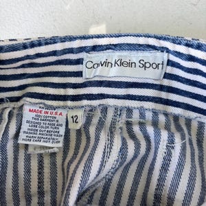 Puede incluir: Primer plano de unos pantalones cortos vaqueros a rayas azules y blancas con una etiqueta "Calvin Klein Sport". Los pantalones cortos tienen una etiqueta de talla 12 y una etiqueta "Made in U.S.A." con instrucciones de cuidado.