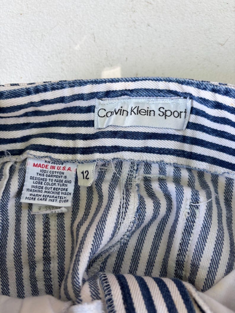 Puede incluir: Primer plano de unos pantalones cortos vaqueros a rayas azules y blancas con una etiqueta "Calvin Klein Sport". Los pantalones cortos tienen una etiqueta de talla 12 y una etiqueta "Made in U.S.A." con instrucciones de cuidado.