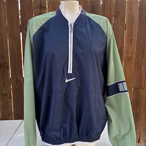 May include: A navy blue and green Nike windbreaker jacket. The jacket has a white zipper, a white Nike logo, and the word "NIKE" on the sleeve. The sleeves are green, and the body is navy blue.