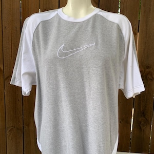 Puede incluir: Camiseta gris y blanca de manga corta con el logo de Nike bordado en el pecho. La camiseta tiene cuello redondo y mangas y paneles laterales blancos. El logo de Nike est&aacute; cosido en blanco.